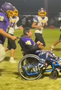 Photograph of a youth football game showing a boy in a wheelchair smiling while being pushed by a player in a purple uniform, with other players in white and yellow uniforms running alongside.
